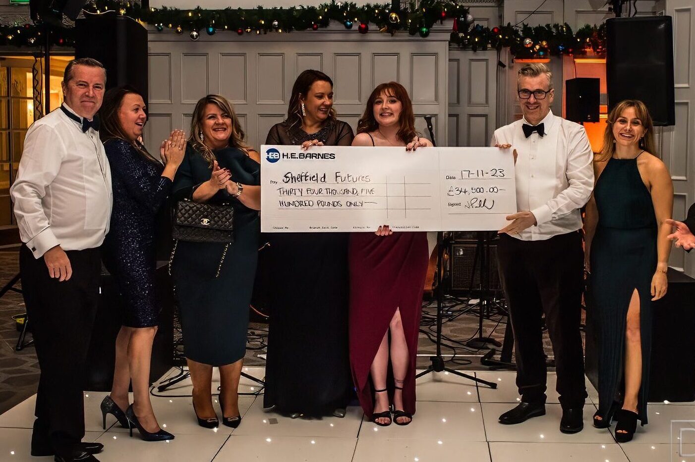 A photograph captures the moment the cheque was handed over. In this image, representatives from HE Barnes and Sheffield Futures are standing on the dancefloor smiling at the camera, holding the giant cheque.